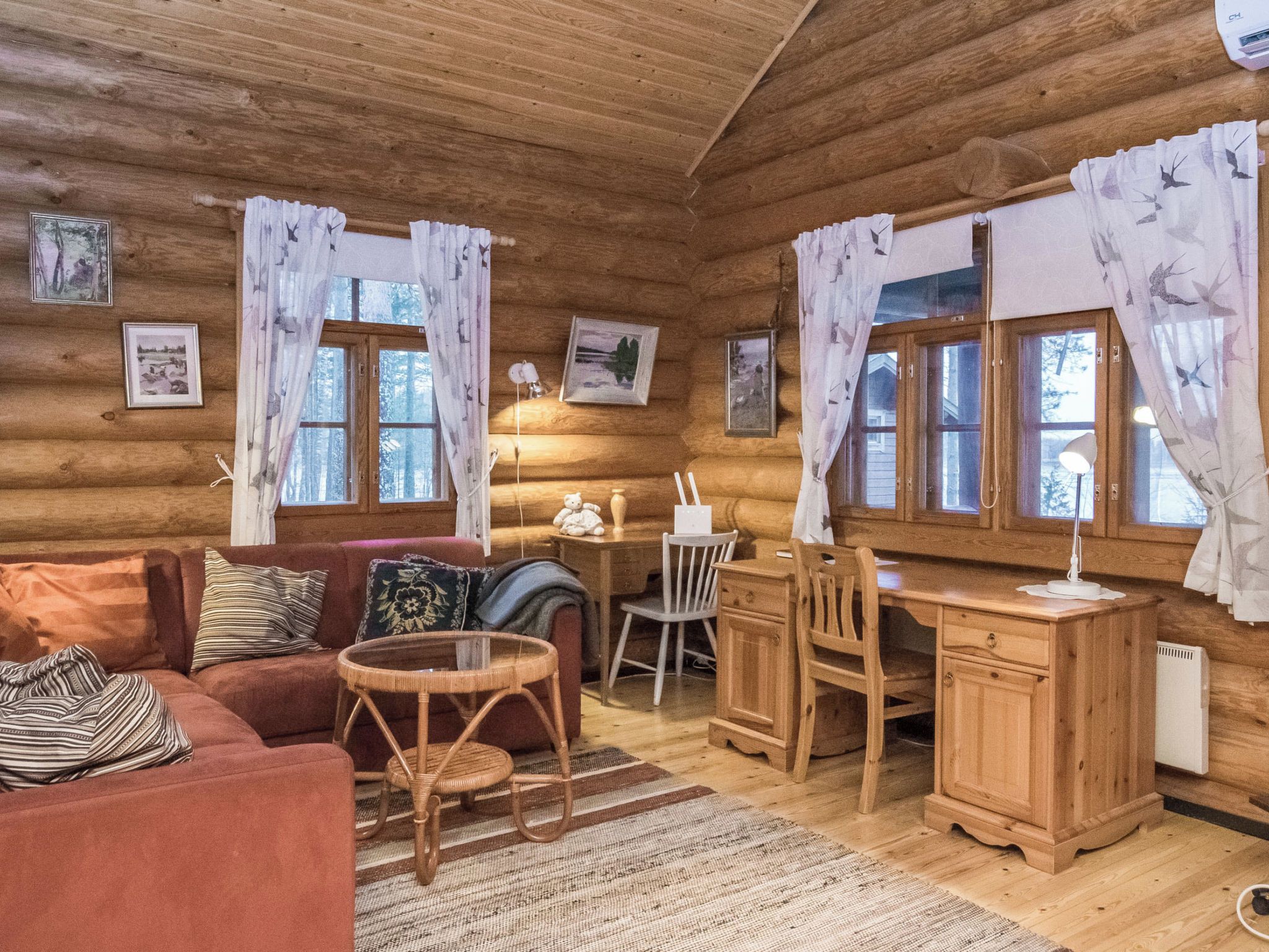 Ein gemütliches Blockhäuschen-Interieur mit Holzvertäfelung, einer Couch, einem Esstisch, Stühlen, einem kleinen Teppich und gerahmten Bildern an den Wänden.