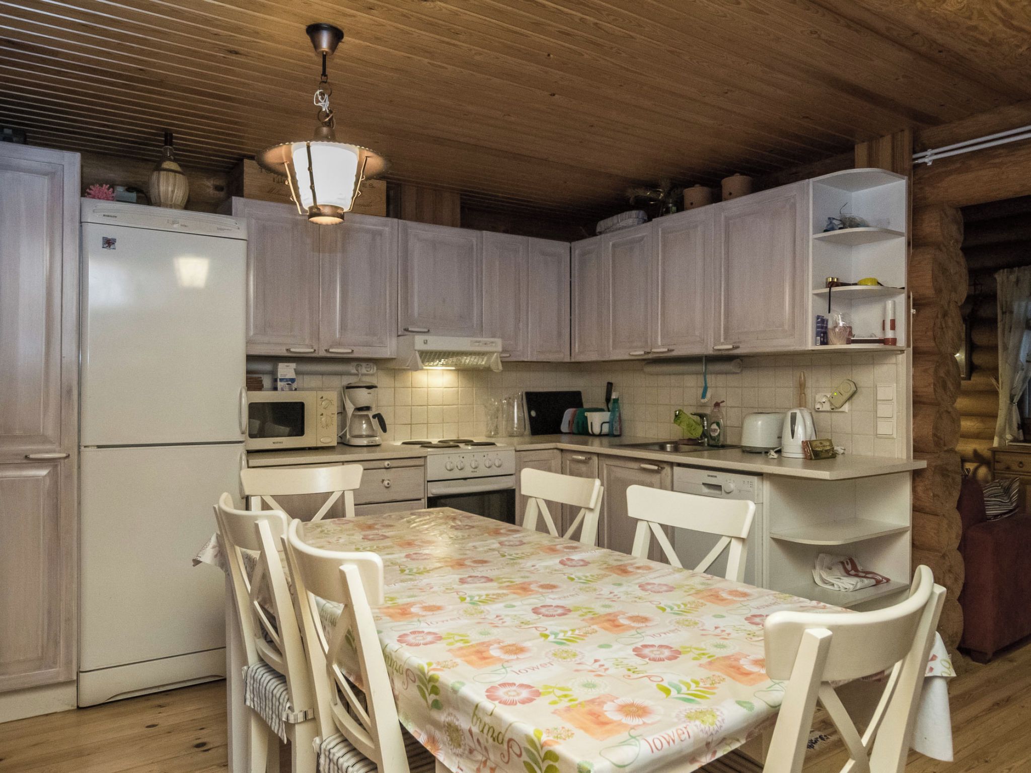 Cozinha compacta com armários brancos, uma mesa de jantar com toalha florida e um lustre.