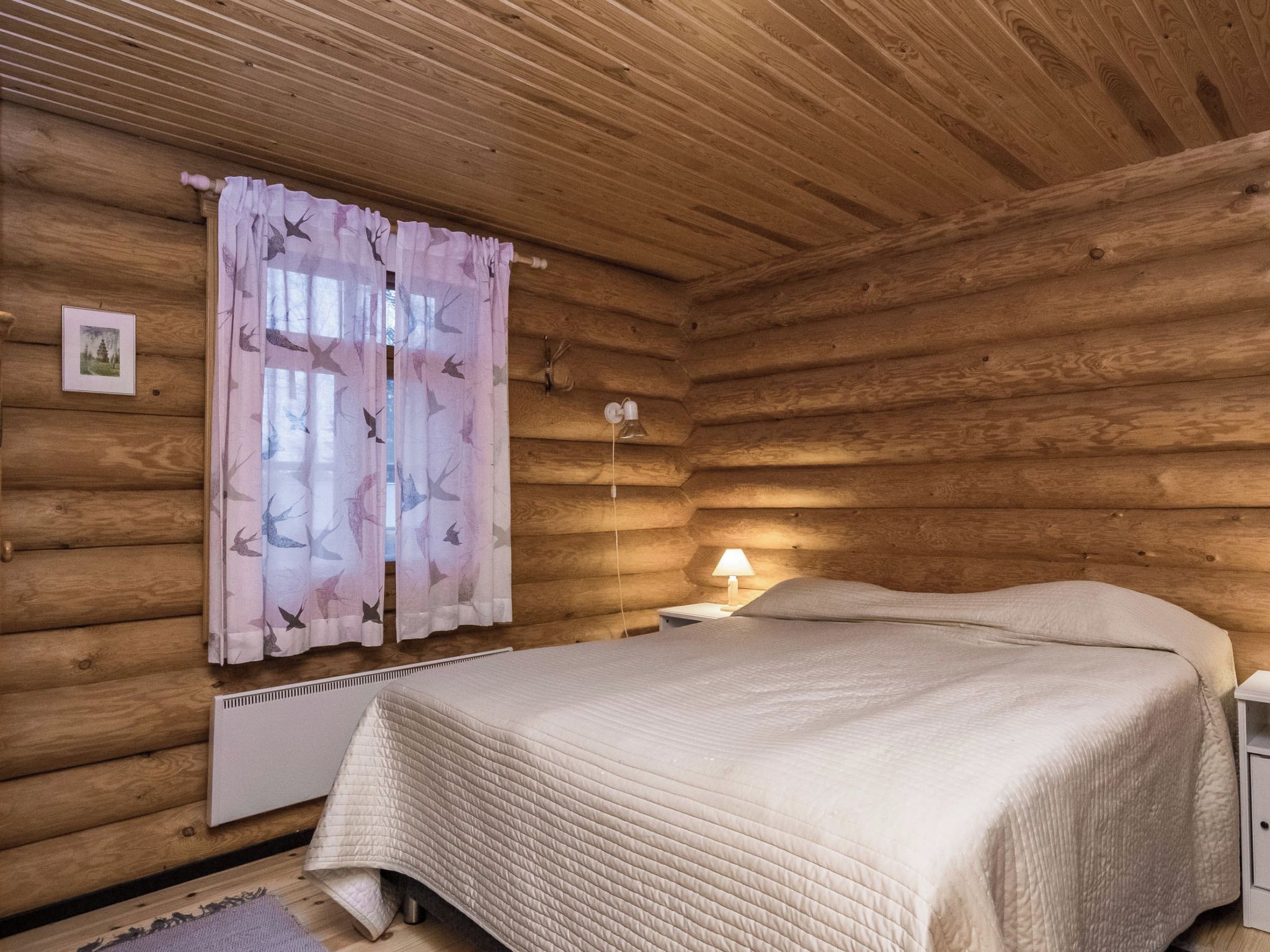Quarto de cabana de madeira com uma cama, mesa de cabeceira, abajur e janela com cortinas.