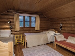 Mökki, jossa on parvelle rakennettu makuuhuone, puulattia, päätyä sisältävä sänky, pieni puutuoli, yöpöytä ja ikkuna, jossa on verhot.