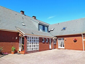 Un bâtiment en briques de deux étages, avec un toit en tuiles, une porte de garage et un petit jardin agrémenté de plantes en pot.