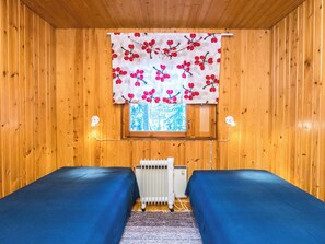 A cabin interior with wooden walls, a window with a view, two beds, a radiator, and a curtain with a floral pattern.