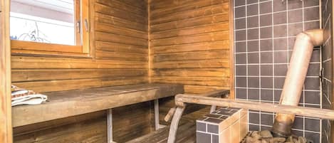 A sauna room with wooden benches, a window, tiled walls, and a wood-burning stove.