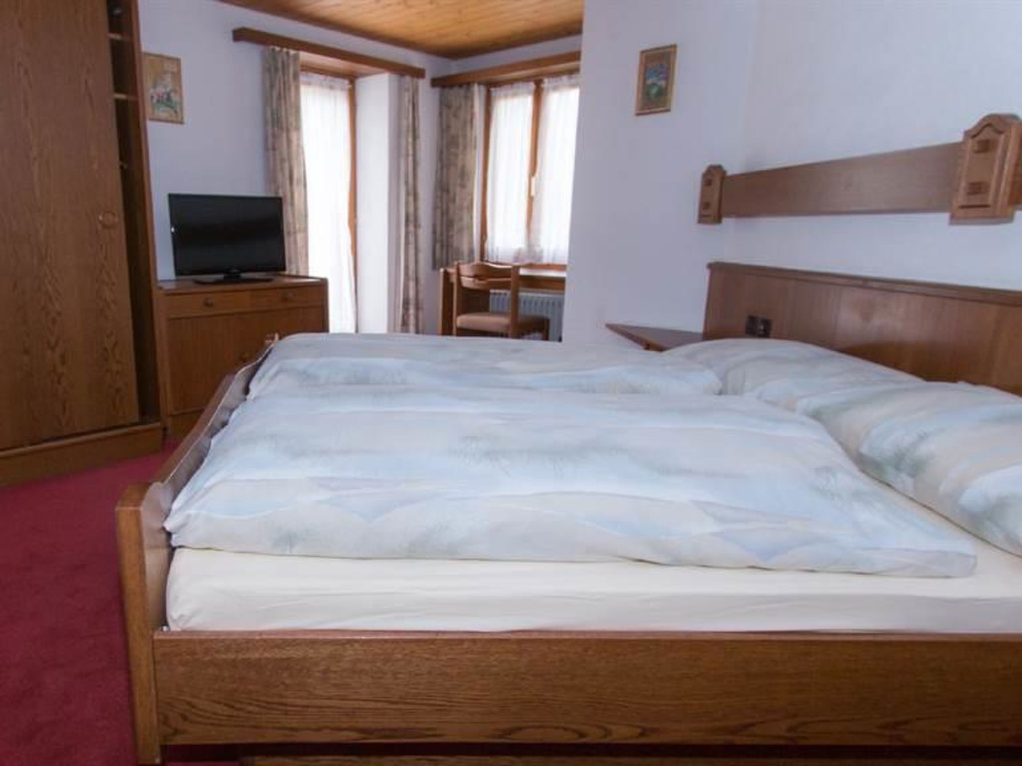 A bedroom with a wooden bed, a television on a wooden cabinet, and a window with curtains.
