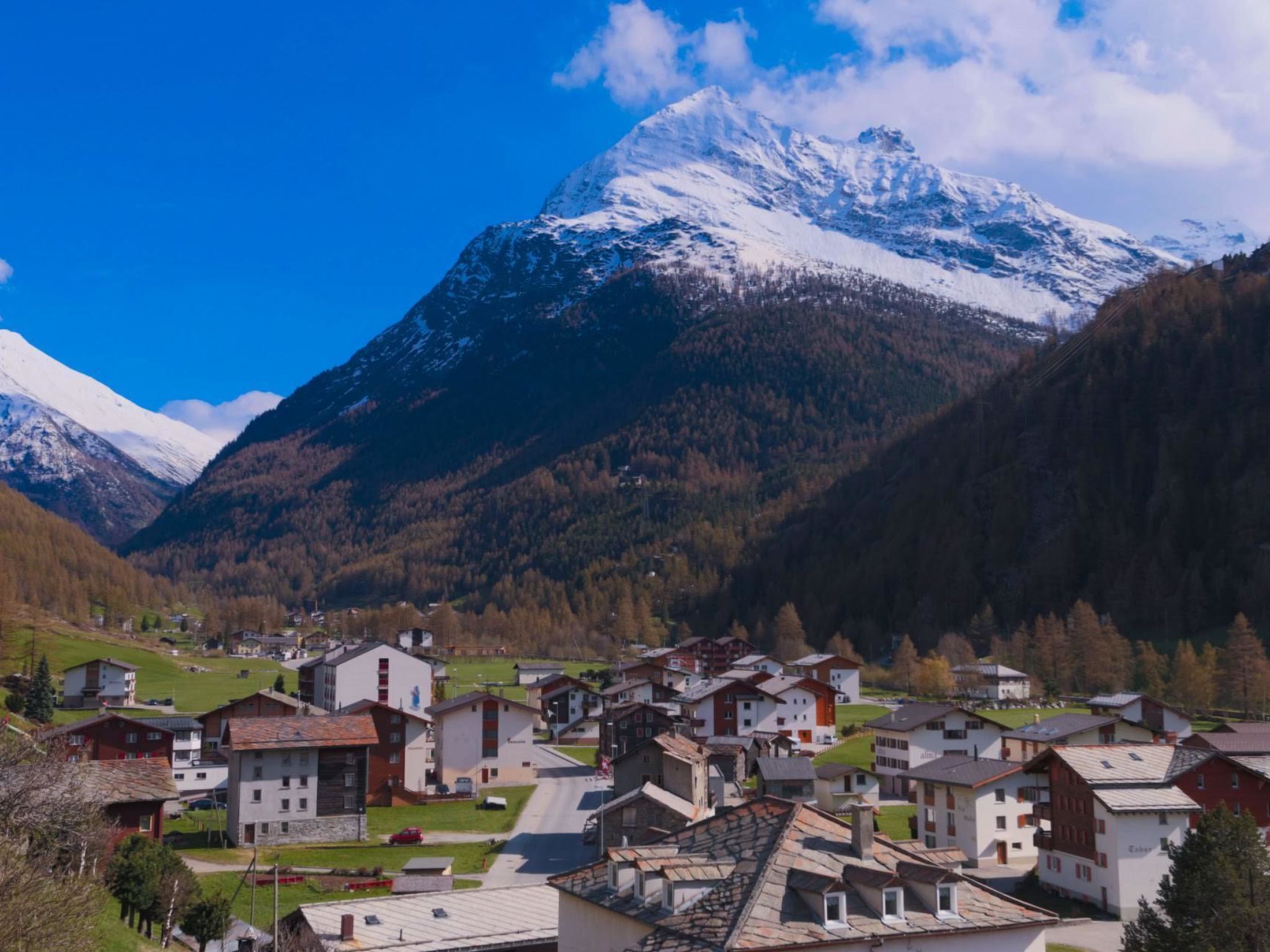 A mountain village with traditional buildings, a clear sky, and a snow-capped peak in the background.