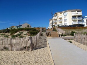 A coastal walkway leading to a beachfront building with multiple balconies and a sandy beach in the background.