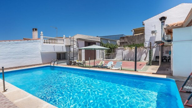 Une piscine rectangulaire à l’eau d’un bleu cristallin, entourée d’un mur blanc et d’une terrasse carrelée. On y trouve des chaises longues et une petite table sous un espace ombragé.