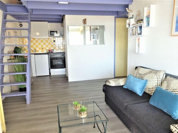 A compact living space with a kitchenette, a sofa with patterned cushions, a glass coffee table, and a ladder leading to a loft area.