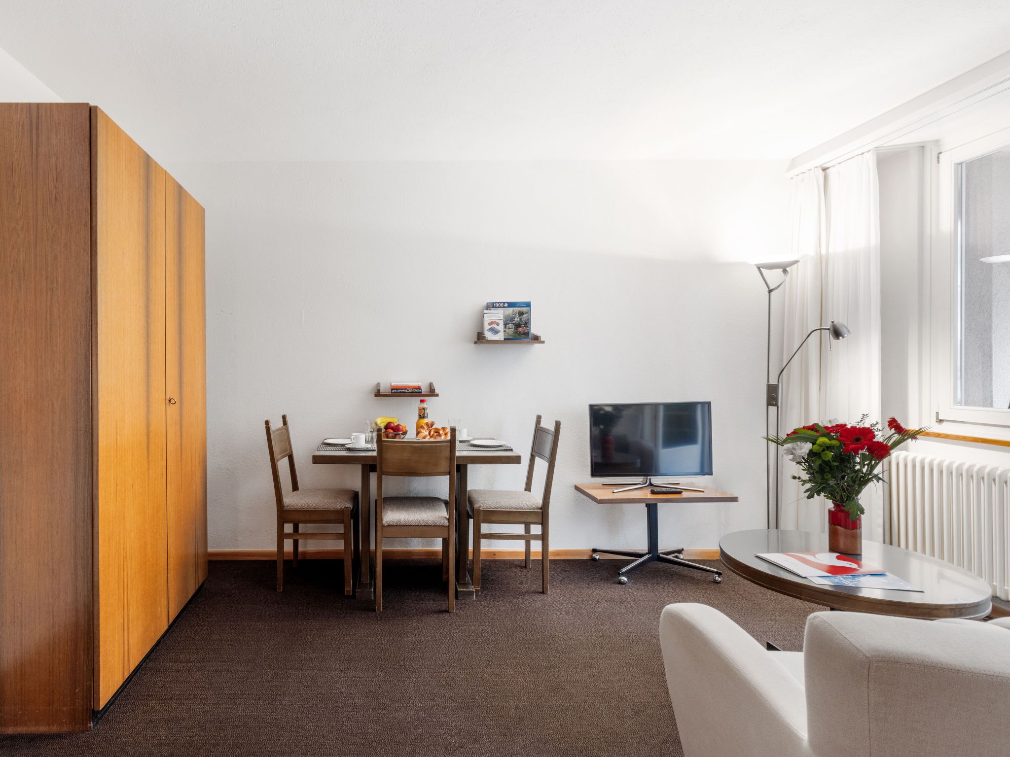 A compact living space with a dining table, chairs, a TV, a small side table, and a radiator.