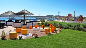 Eine Strandlounge mit Sitzgelegenheiten und Sonnenschirmen, ein Spielbereich für Kinder sowie ein Blick auf das Meer und die dahinterliegenden Hügel.