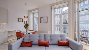 Een moderne woonkamer met een grijze bank, rode kussens, een eethoek met een tafel en stoelen, en grote ramen met uitzicht op de stad.