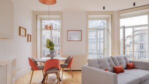 Een moderne woonkamer met een grijze bank, rode en grijze stoelen, een ronde tafel met een vaas, en grote ramen met uitzicht op de stad.