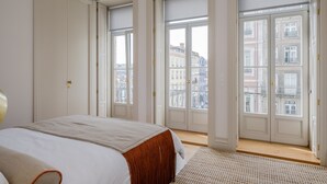 Een slaapkamer met een groot bed, uitzicht op de stad door grote ramen en een deur die naar een andere kamer leidt.