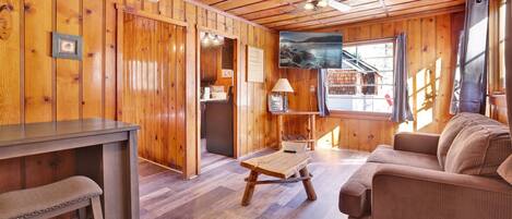 A cozy cabin interior with wooden walls, a dining area, a sofa, and a large window with a view of a lake.