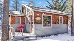 A small cabin with a red chair outside, a wooden sign reading "Little Pines," and a snow-covered ground. - Little Pines #102 (Big Bear Lake)