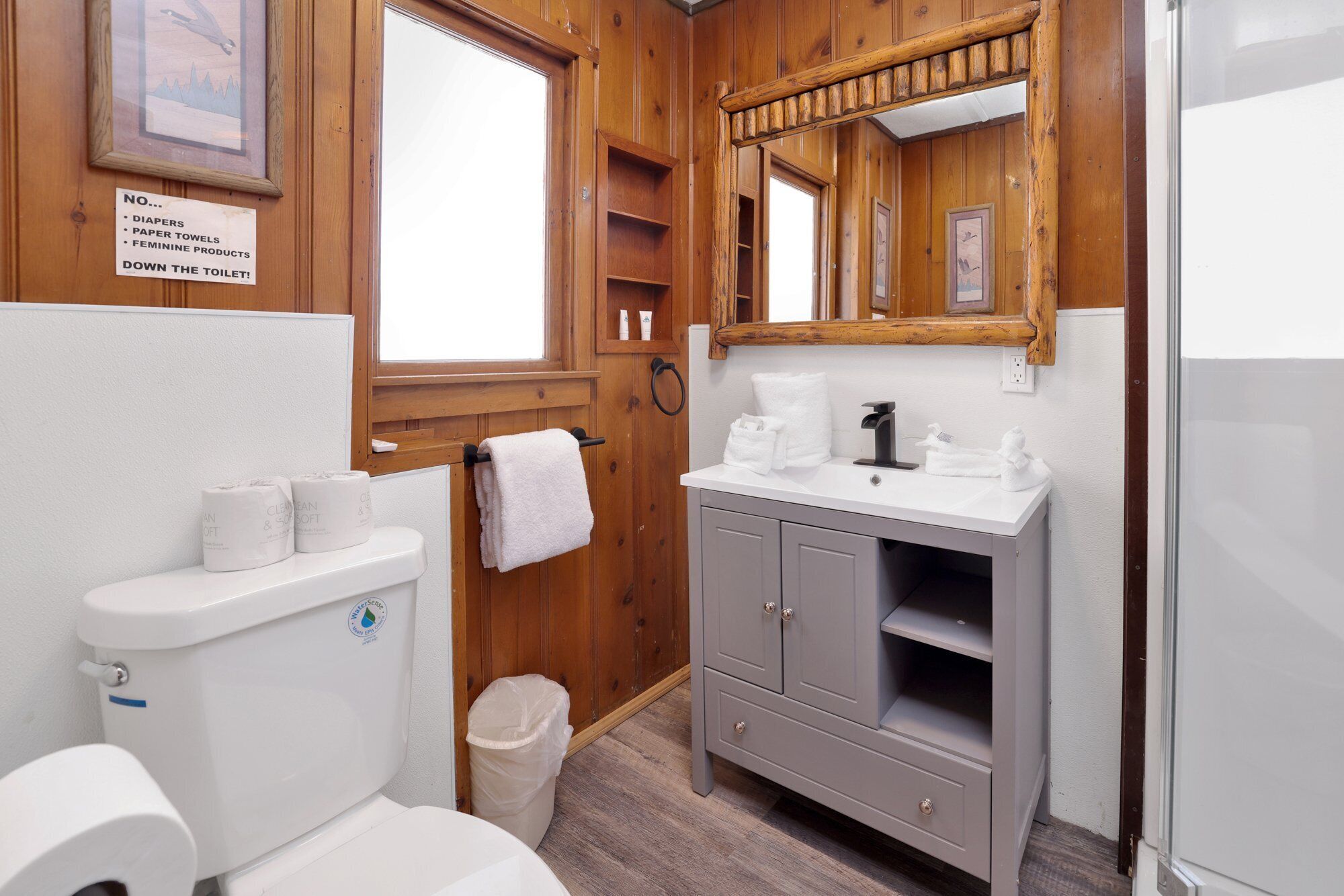 Une salle de bain équipée de toilettes, d’un lavabo et d’un miroir. Un panneau indique qu’il est interdit de jeter les couches, les essuie-tout et les produits d’hygiène féminine dans les toilettes.