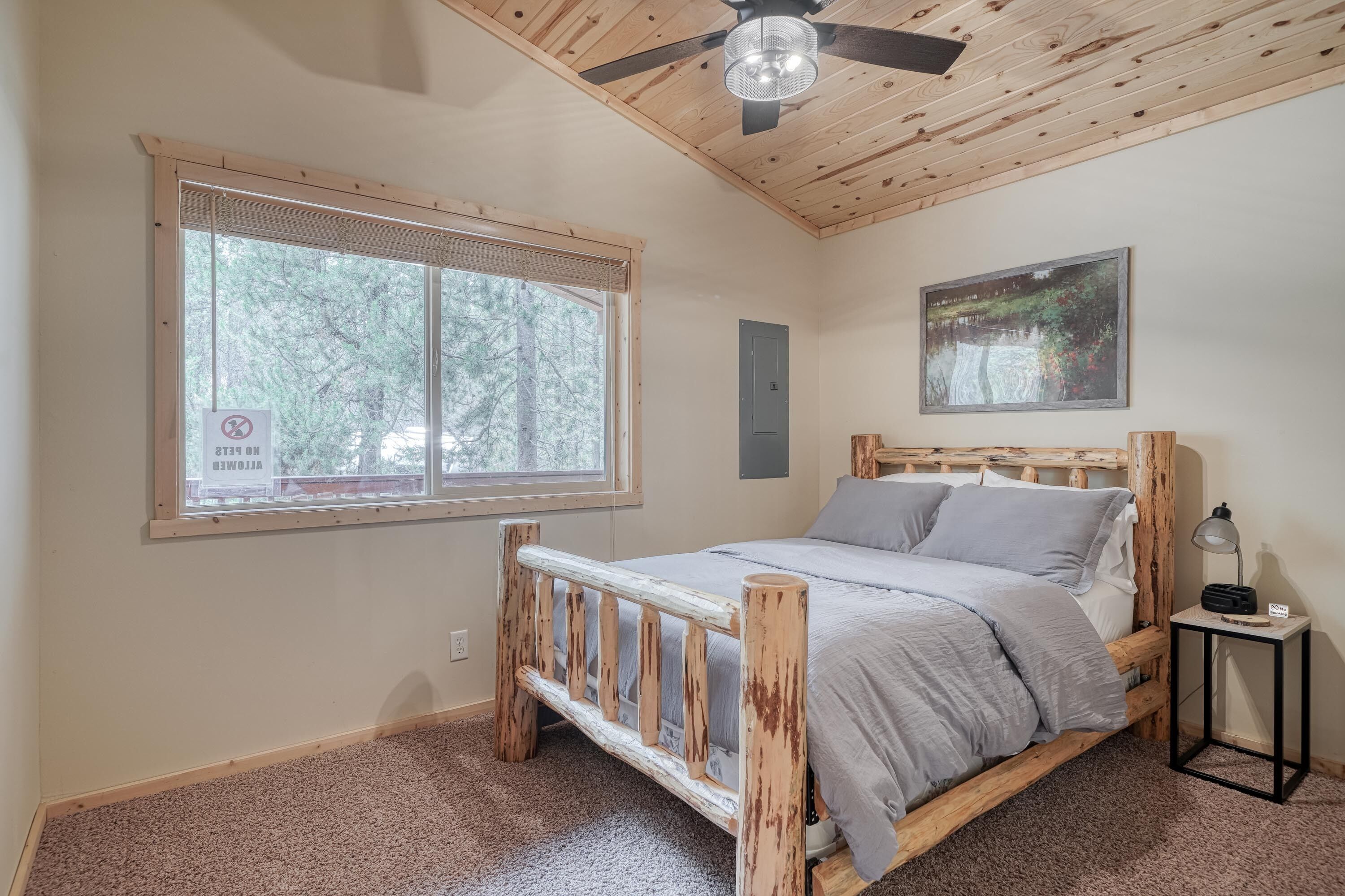 A bedroom with a wooden bed, a window with a view of trees, a ceiling fan, and a small bedside table with a lamp.
