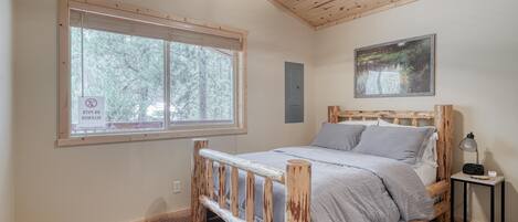 A bedroom with a wooden bed, a window with a view of trees, a ceiling fan, and a small bedside table with a lamp.