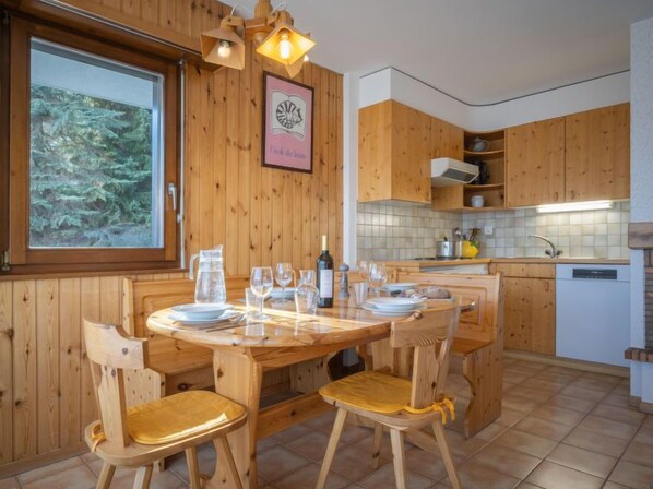 Un interior de madera con una mesa de comedor dispuesta para cuatro, armarios de madera, una ventana con vista a árboles y una zona de cocina con un lavamanos y encimera.