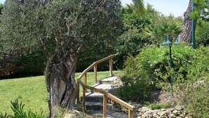 Un escalier en pierre mène à une terrasse en bois, elle-même entourée d’une végétation luxuriante et d’un grand arbre.