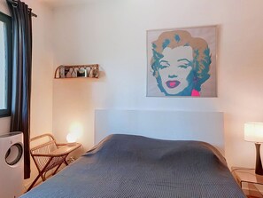 Une chambre d’hôtel avec un lit, une chaise, une étagère murale avec des livres, une lampe et une grande affiche de style pop art.