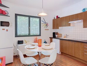 Une cuisine moderne avec des meubles blancs, une table à manger pour quatre personnes et une fenêtre donnant sur un paysage verdoyant.