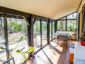 Un espace extérieur abrité, doté d’une table et de chaises, de grandes fenêtres et offrant une vue sur la verdure environnante.