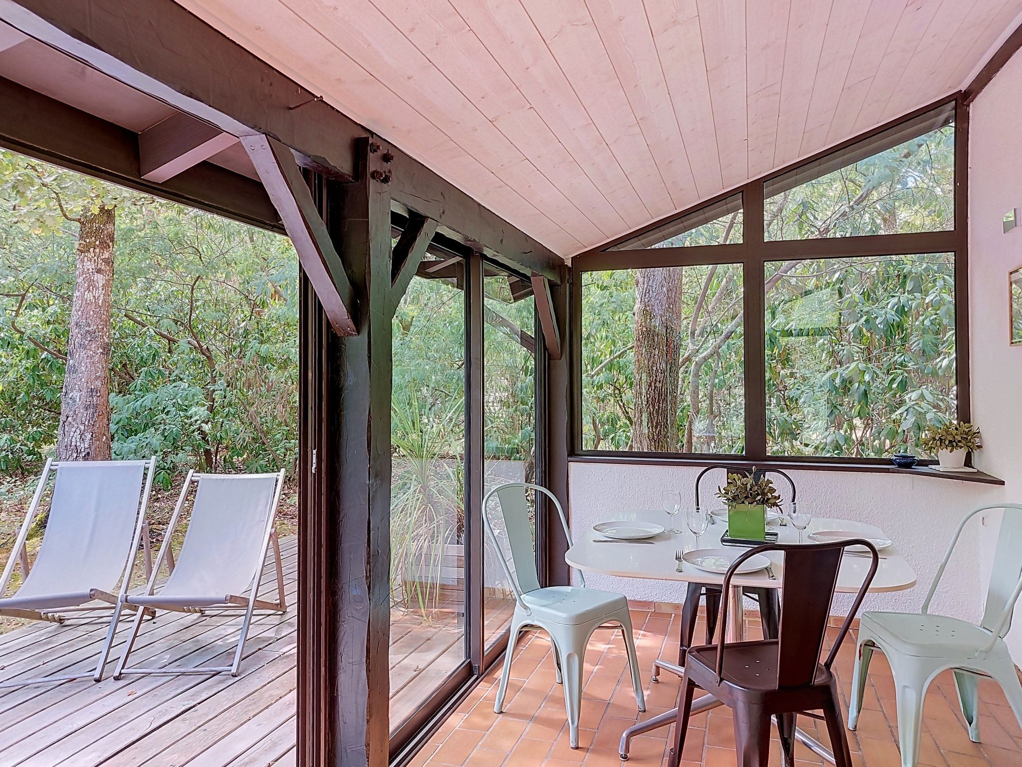 A covered outdoor seating area with a table and chairs, surrounded by large windows offering views of trees and foliage.