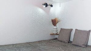 A corner of a room with a textured white wall, a stone floor, a white vase with dried plants, and two patterned cushions. - Cimes de Caron 1603 by Interhome (Val Thorens)