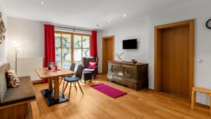 Un salon chaleureux avec une table à manger en bois, un canapé, un téléviseur fixé au mur et une grande fenêtre ornée de rideaux rouges.