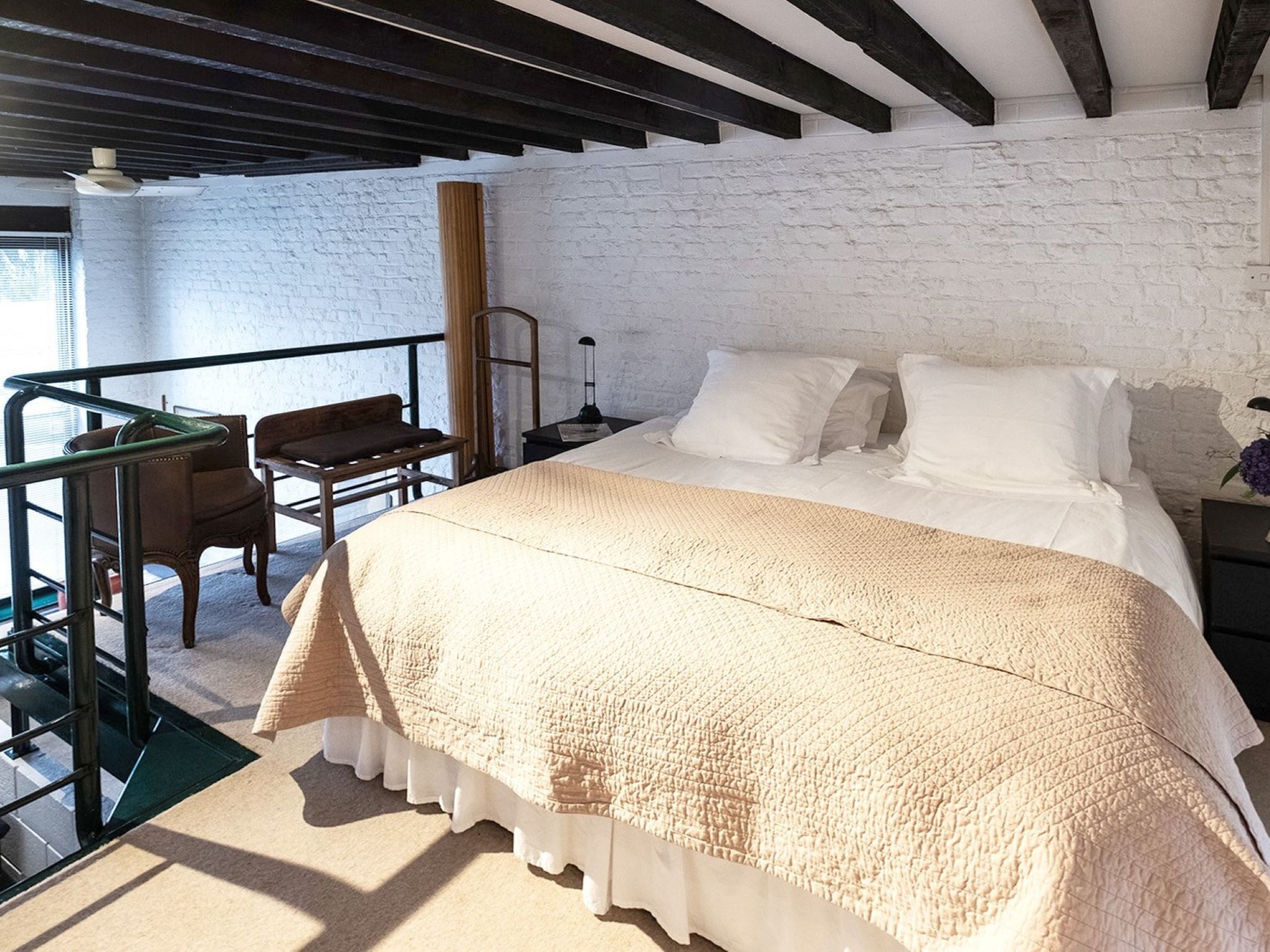 Un dormitorio con una cama grande, una silla de madera, una mesita y una pared de ladrillo blanco.