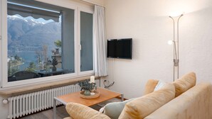 A living room with a sofa, a small table with a candle and a vase, a TV mounted on the wall, and a view of mountains through a large window. - Double Room Classic by Interhome (Ascona)