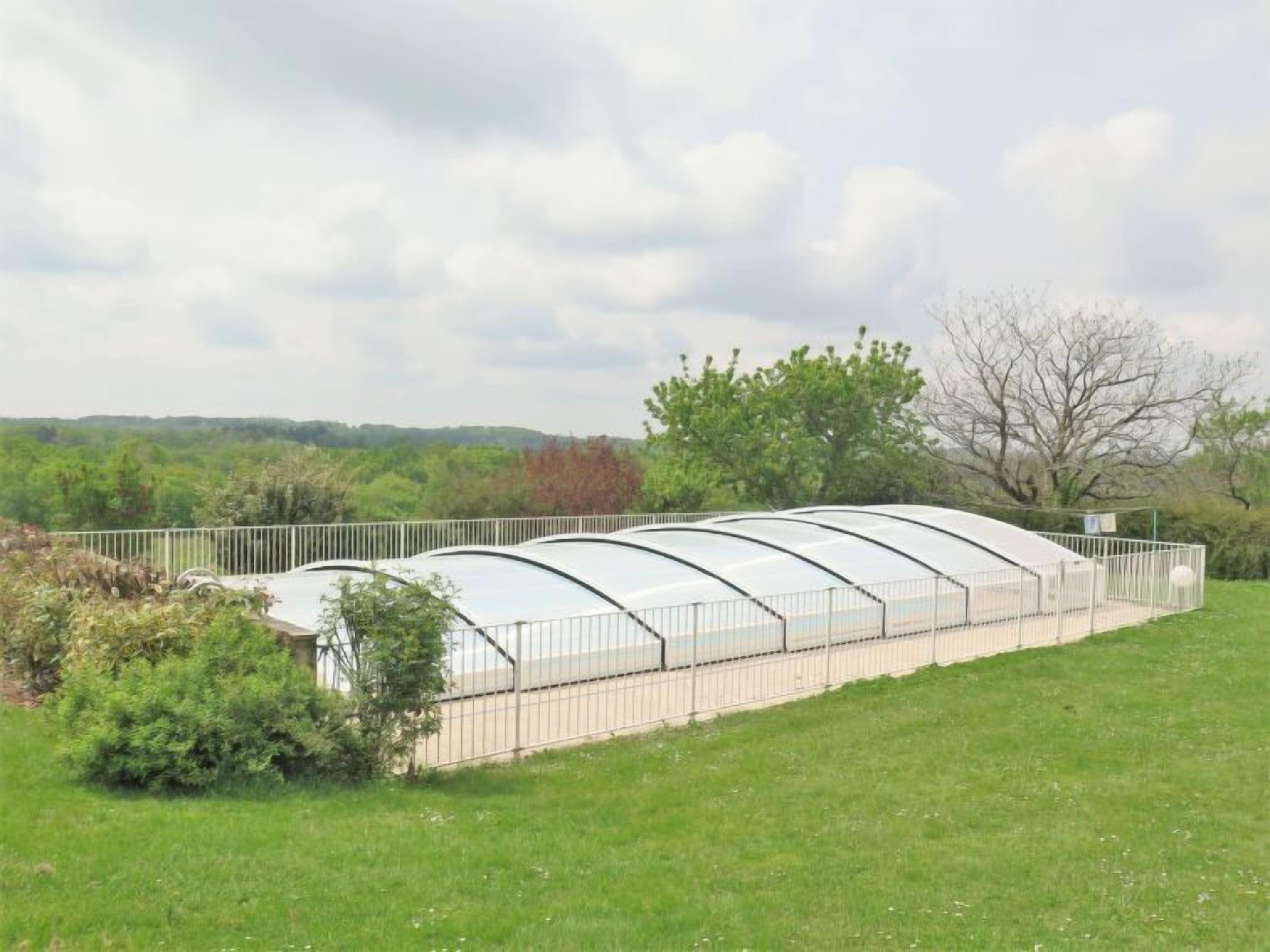 A greenhouse structure with a curved roof, surrounded by a metal fence, set in a grassy area with trees in the background.