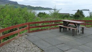 Une table de pique-nique avec des bancs, installée sur une plateforme en béton et entourée d’une clôture en bois, d’où l’on peut admirer un plan d’eau et des collines lointaines.