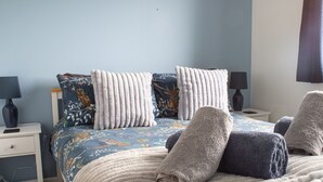 Un lit bien fait, recouvert d’une couvre-lit à motifs floraux bleus, avec deux oreillers blancs et deux serviettes. Il y a deux tables de chevet avec des lampes et un petit tiroir.