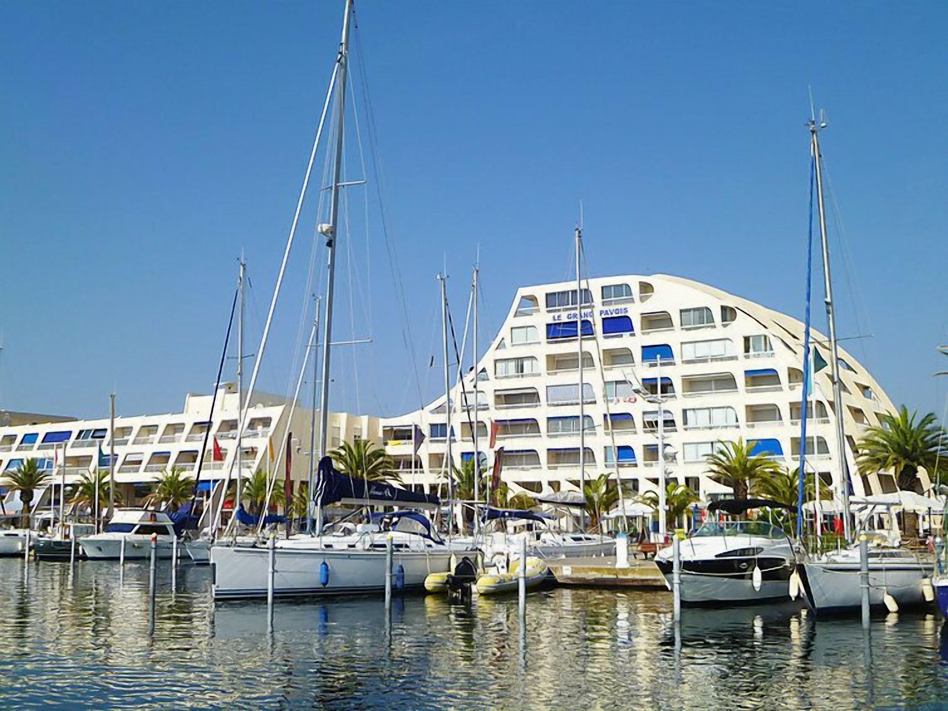 A marina with multiple boats docked, a large building with balconies, and palm trees.
