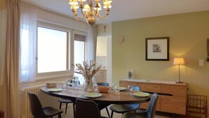 A dining area with a round table, six chairs, a chandelier, a window with curtains, a radiator, a wooden cabinet, and a framed picture on the wall. - Barzettes-Vacances 558 / Entrée N° 18 by Interhome (Crans-Montana)