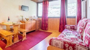 Ein gemütliches Zimmer mit Holzboden, einem Holztisch mit Stuhl, einer gemusterten Couch und einem großen Fenster mit Vorhängen.