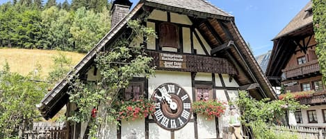 A traditional half-timbered building with a large outdoor clock, surrounded by greenery and a clear sky.