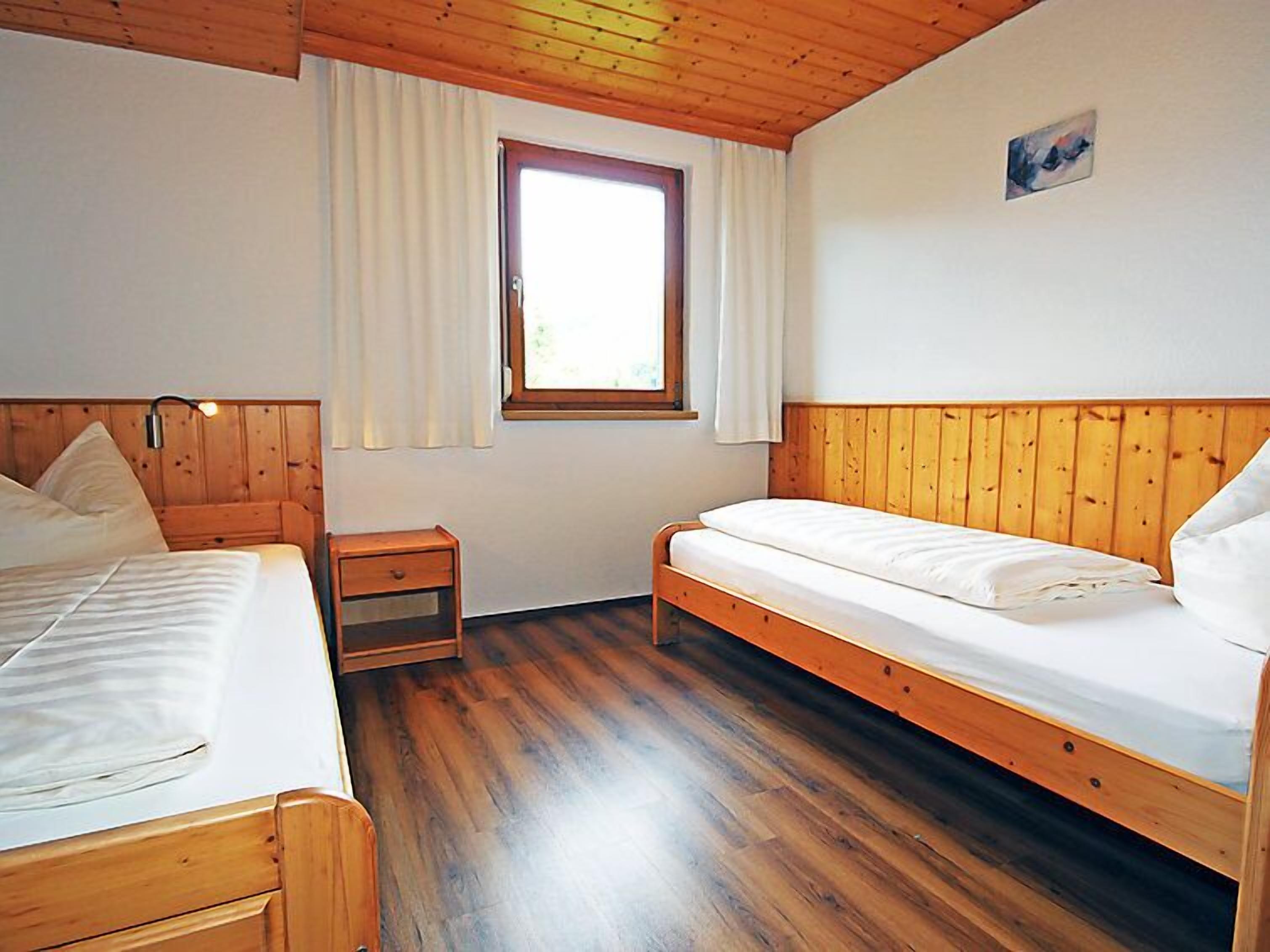 A room with two wooden beds, a small bedside table, a window with curtains, and a painting on the wall.
