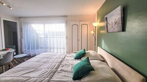 A hotel room with a bed, a desk, and a chair. There is a window with sheer curtains and a door in the background. - Les Embruns by Interhome (Carnac)