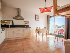 A modern kitchen with white cabinets, a black countertop, and a stainless steel range hood. There is a dining area with a table and chairs, and a view of the sea through large windows. - Le Grand Large by Interhome (Capbreton)