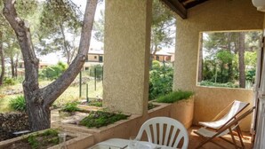 Un espace extérieur abrité, aménagé pour les repas, avec une table dressée pour deux personnes, entouré de chaises blanches, d’un fauteuil et offrant une vue sur un jardin et des bâtiments en arrière-plan.