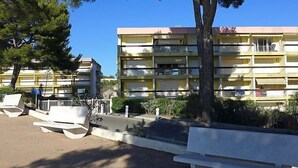 Un immeuble à plusieurs étages doté de balcons, une zone pavée avec des bancs, et un arbre au premier plan.