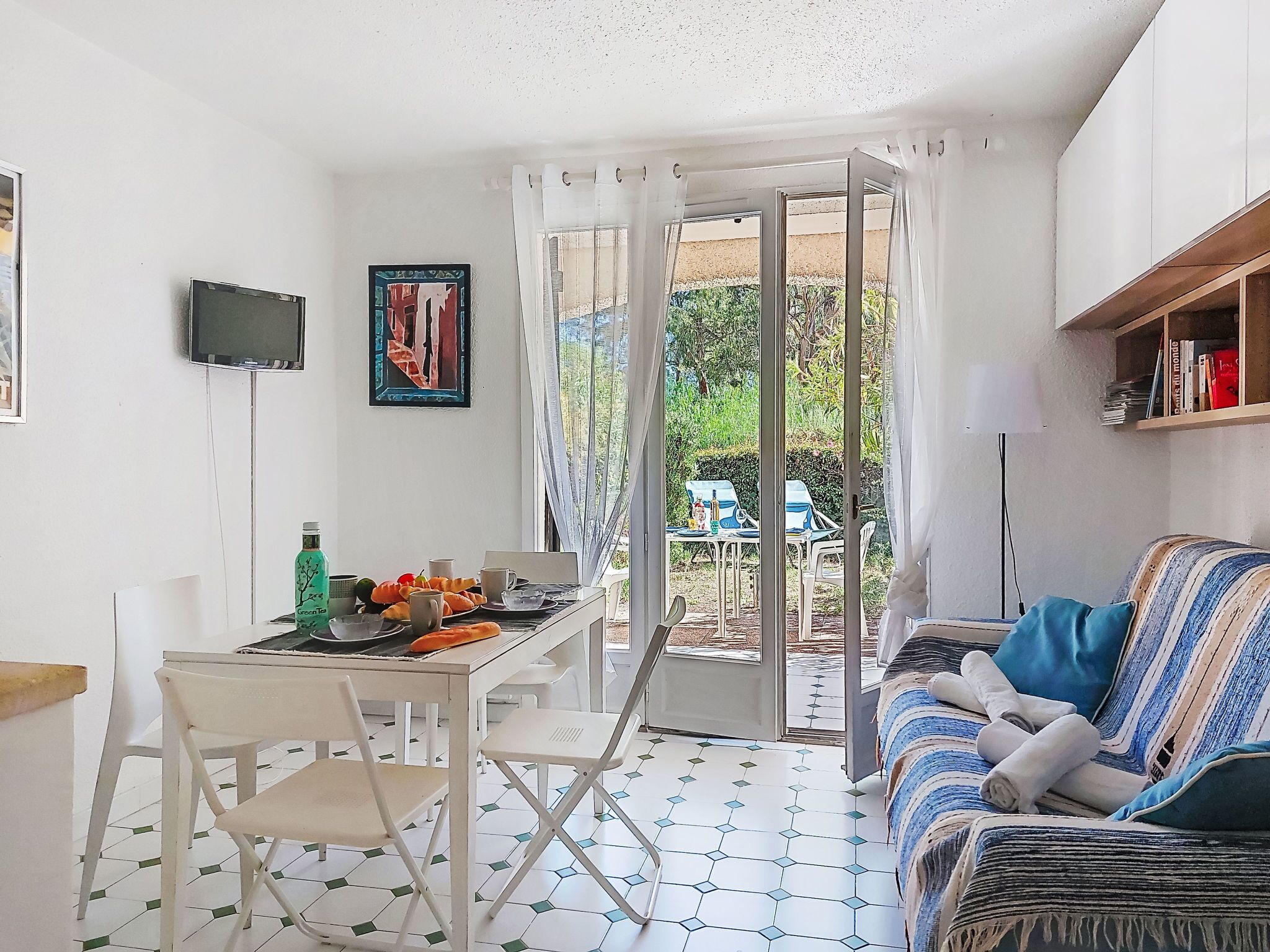 A room with a dining table, chairs, a sofa, and a TV. There is a window with curtains, a bookshelf, and a tiled floor.