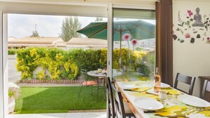 A dining area with a table set for two, a green umbrella, and a view of a garden and buildings outside. - Les Cristallines by Interhome (Cap d'Agde)