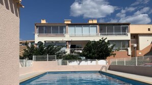 A swimming pool with a tiled deck, surrounded by a fence, and a building with balconies in the background. - Les Cristallines by Interhome (Cap d'Agde)