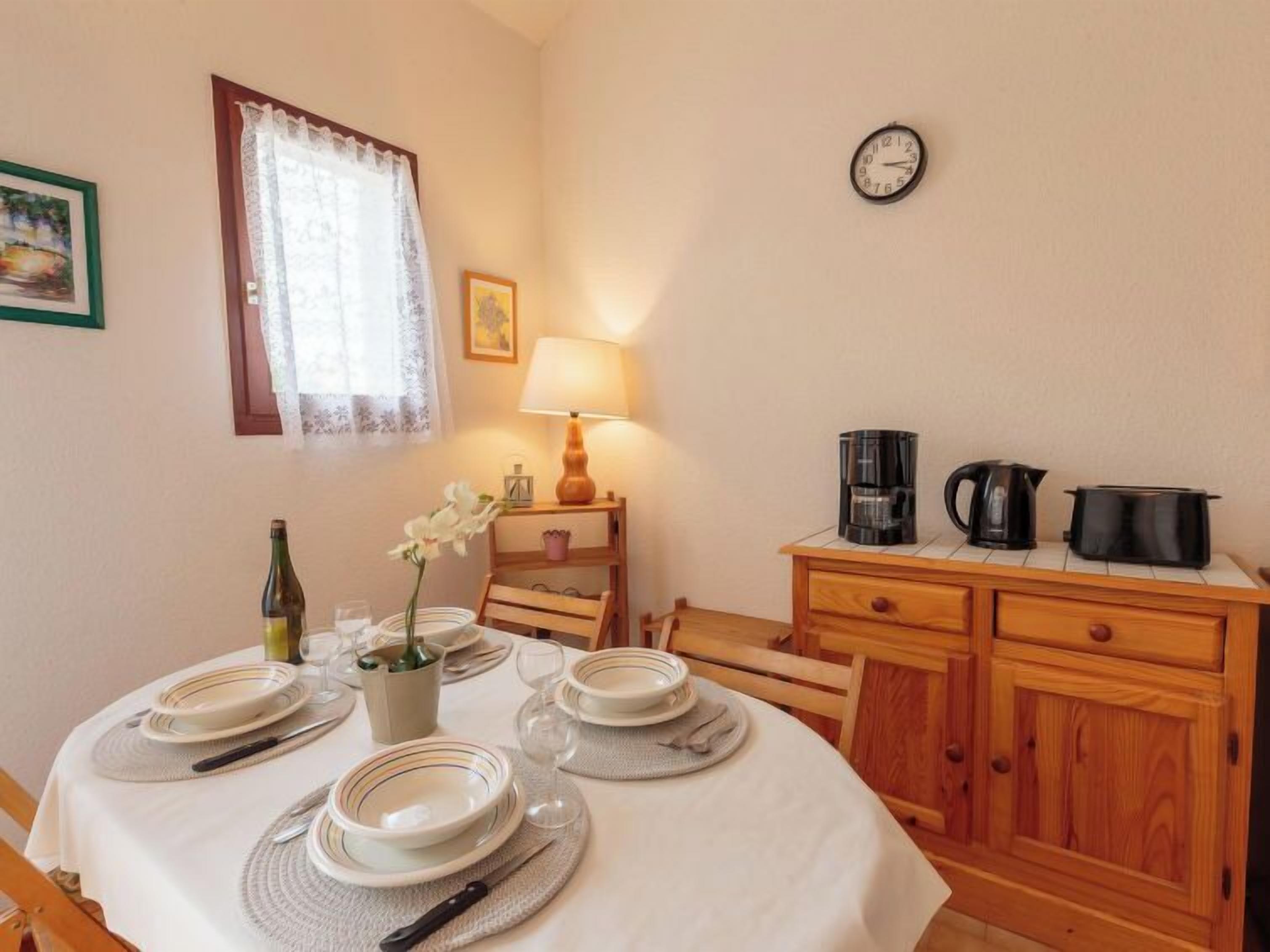 A dining area with a table set for four, a clock on the wall, and a kitchen area with appliances.