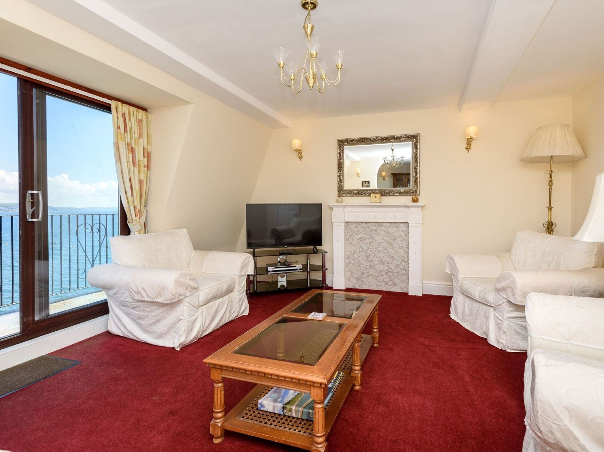 A living room with a red carpet, white sofas, a glass-top coffee table, a fireplace, a TV, and a balcony with curtains.