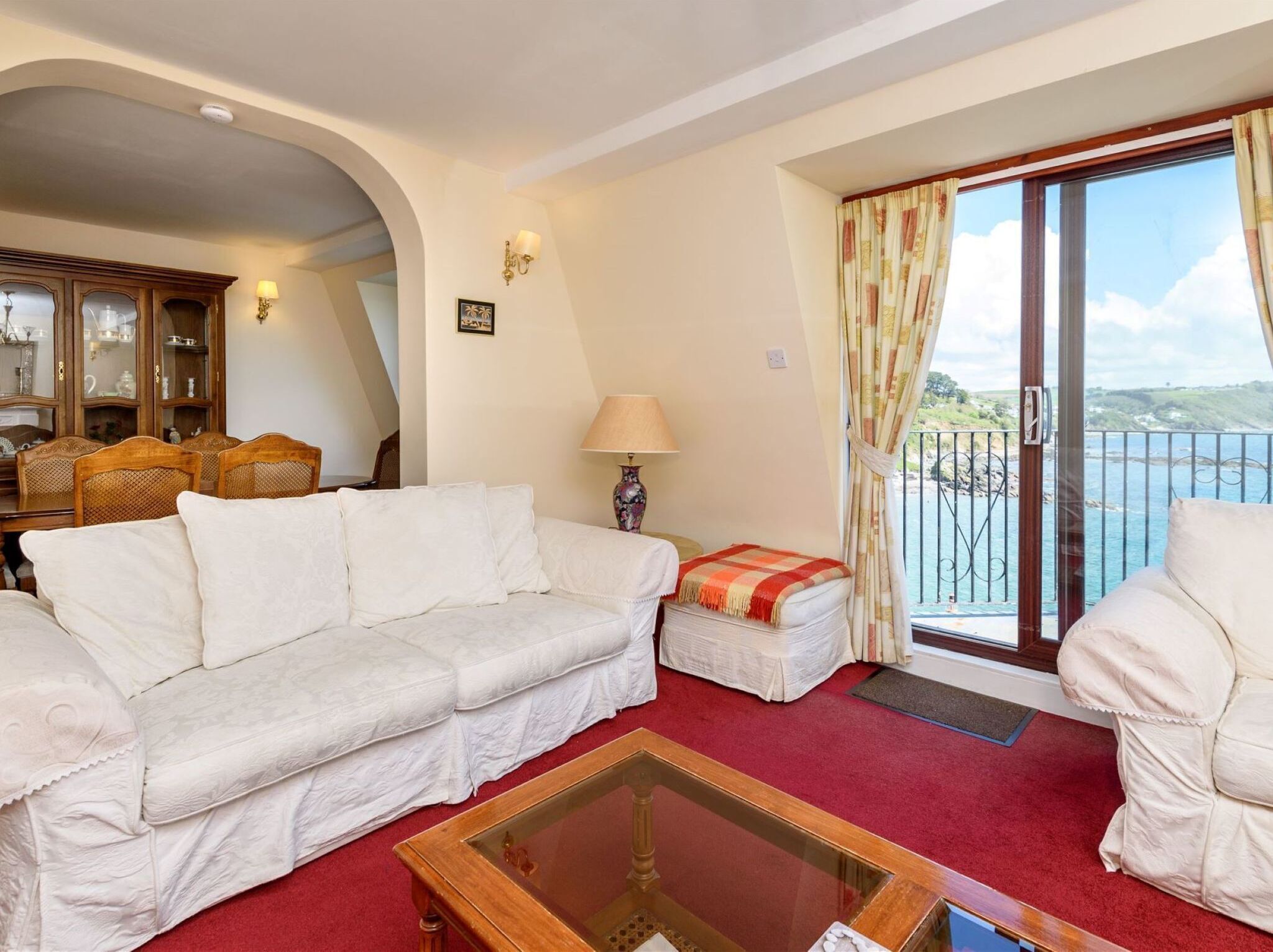 A living room with a white sofa, a glass coffee table, and a view of the sea through a balcony.